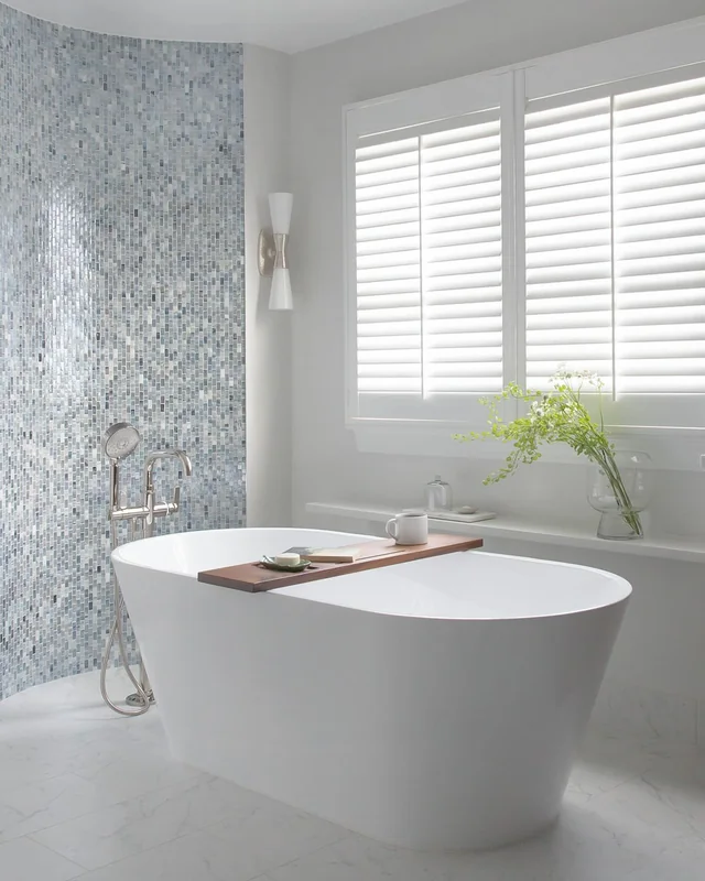 Freestanding tub beside a blue mosaic tile wall and plantation shutters in a remodeled bathroom